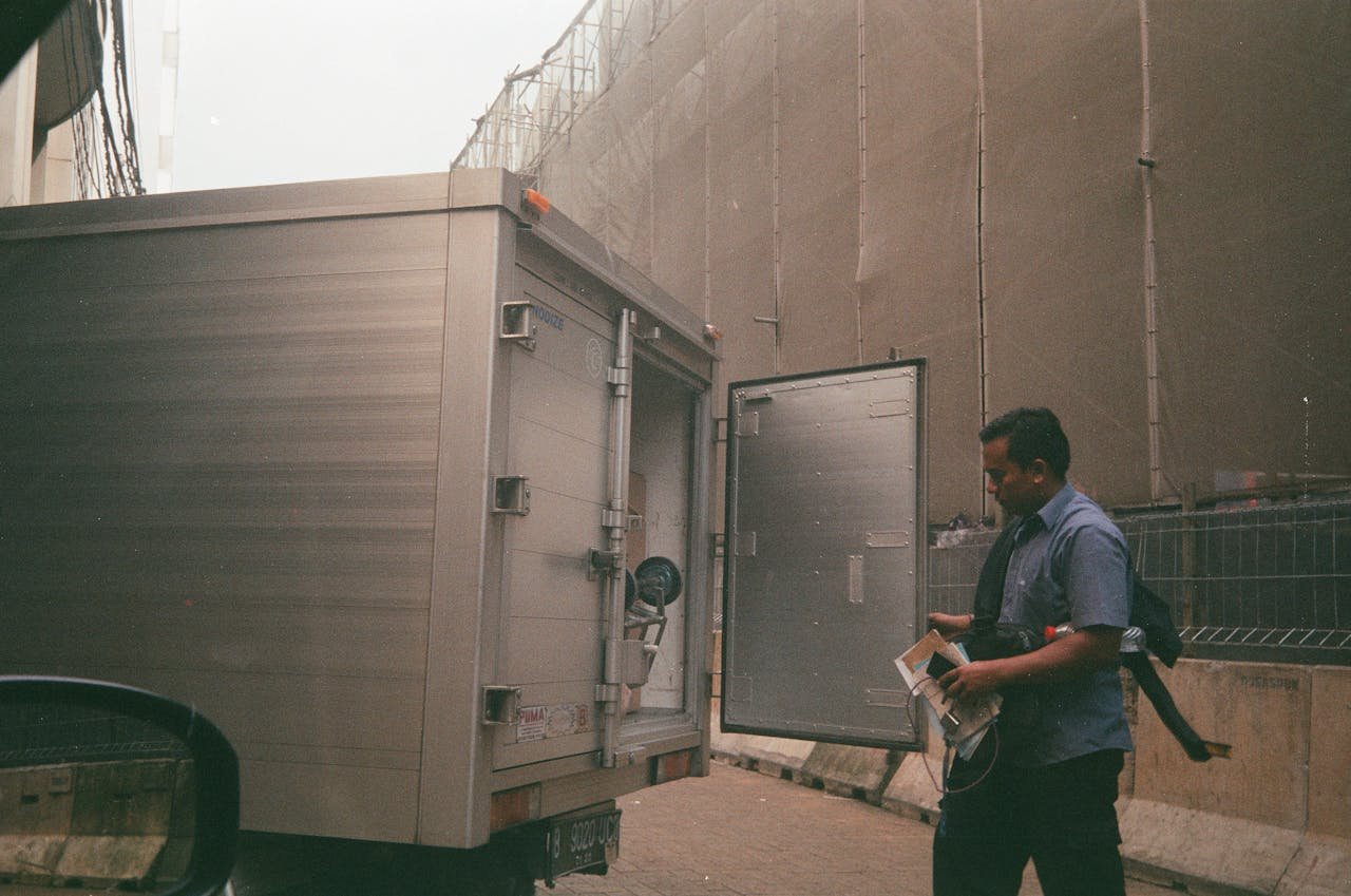 A logistics worker in a blue shirt checking a cargo truck on a city street. Industrial background suggests transportation and delivery.